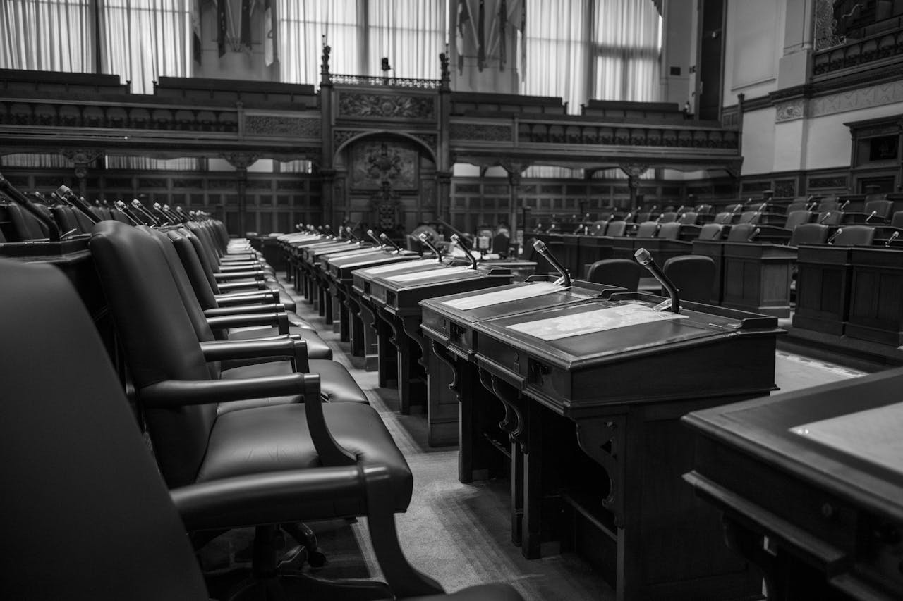 Black and white photo of a traditional legislative assembly chamber, featuring empty seats and desks.